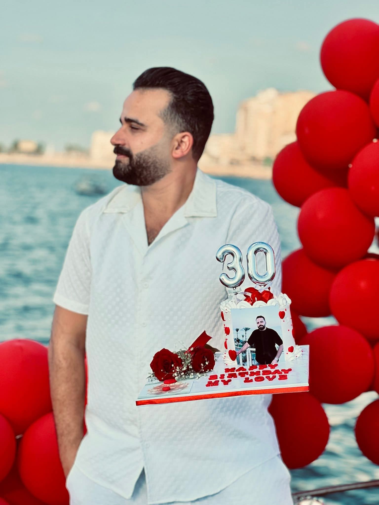 رجل ملتحٍ بقميص أبيض يحمل كعكة عيد ميلاده الثلاثين مع بالونات حمراء وورود أمام البحر.